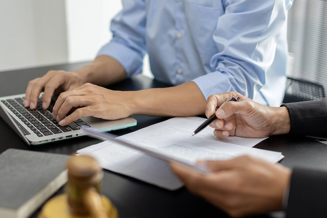 Two people collaborate at a desk, one typing on a laptop and the other writing notes, with legal documents and a gavel nearby.